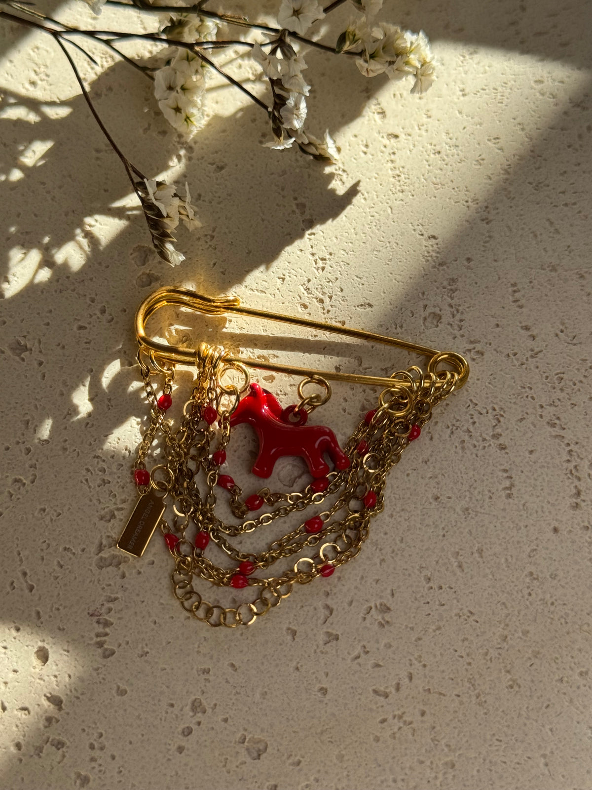 A handmade statement brooch featuring a glossy red horse charm, layered gold-tone chains and small red beads. The rich red and gold tones add a playful accent to a blazer, coat or bag. Size: 5 × 5 cm.